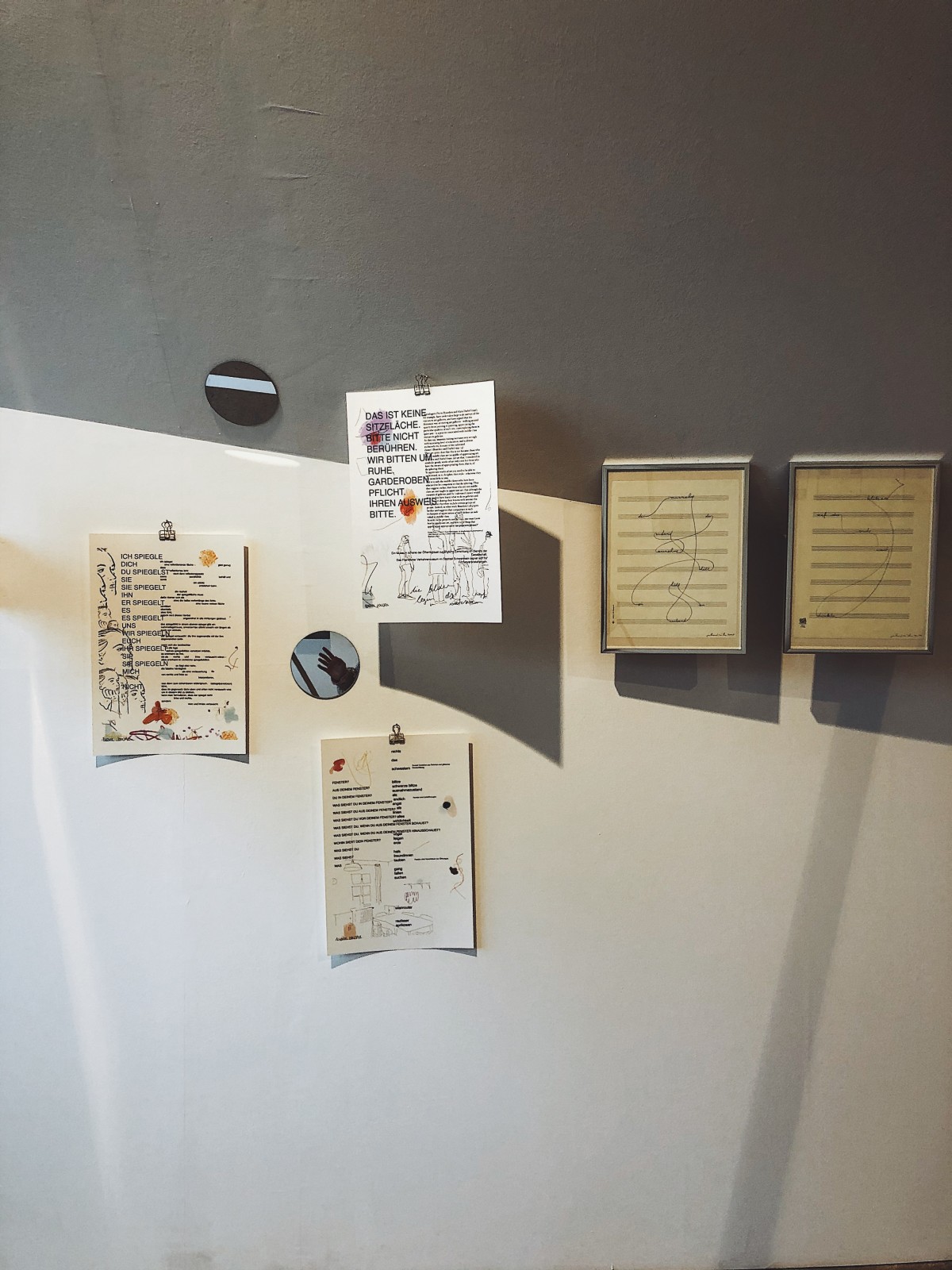 Fünf Text und Illustrationsbildern an einer Wand hängend. Daneben zwei kleine runde Spiegel.